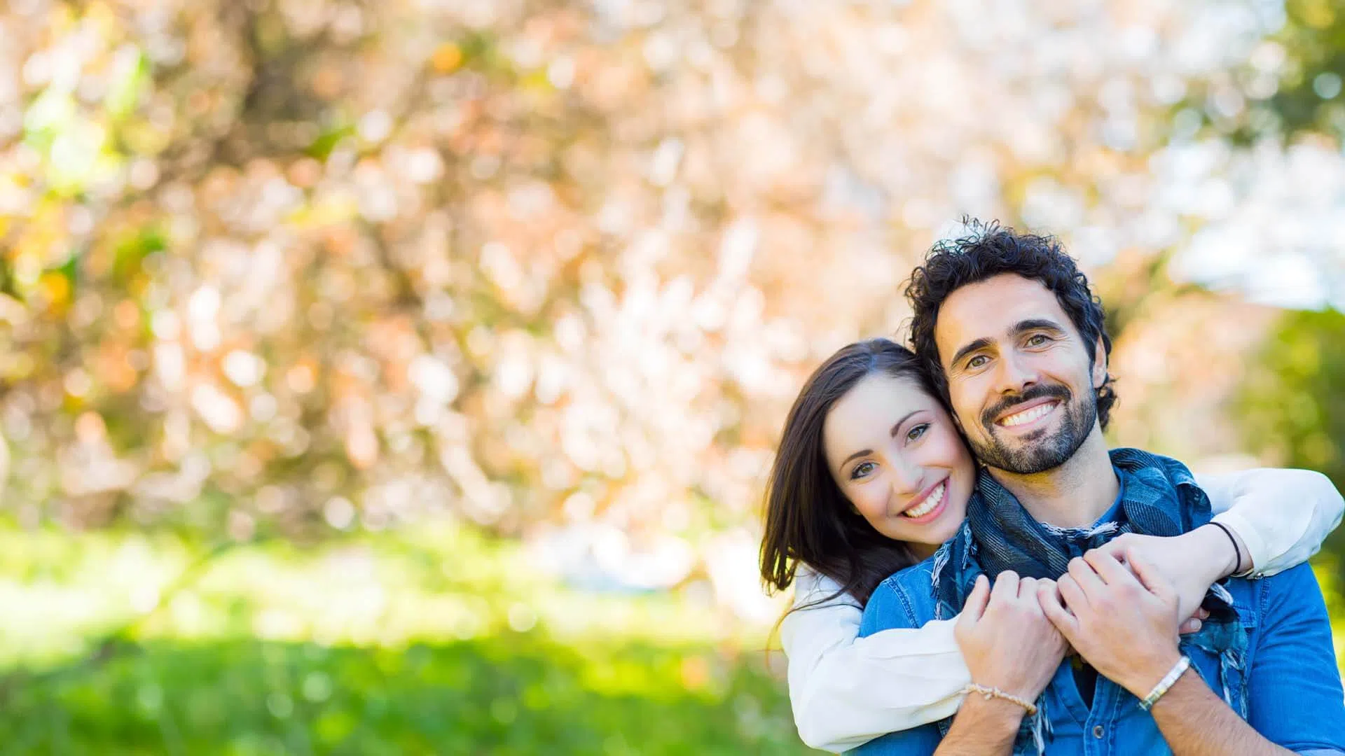 Smiling couple embracing outdoors with blurred greenery and autumn foliage in the background.