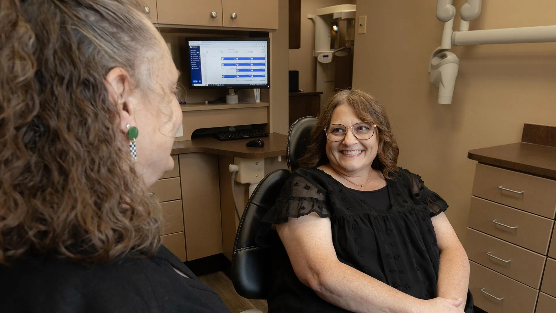 Smiling woman sitting in dental chair talking with another woman in dental office with computer screen.