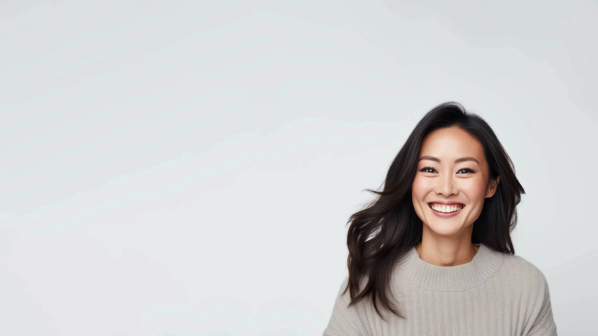 Smiling Asian woman with long hair wearing a beige sweater on a light gray background