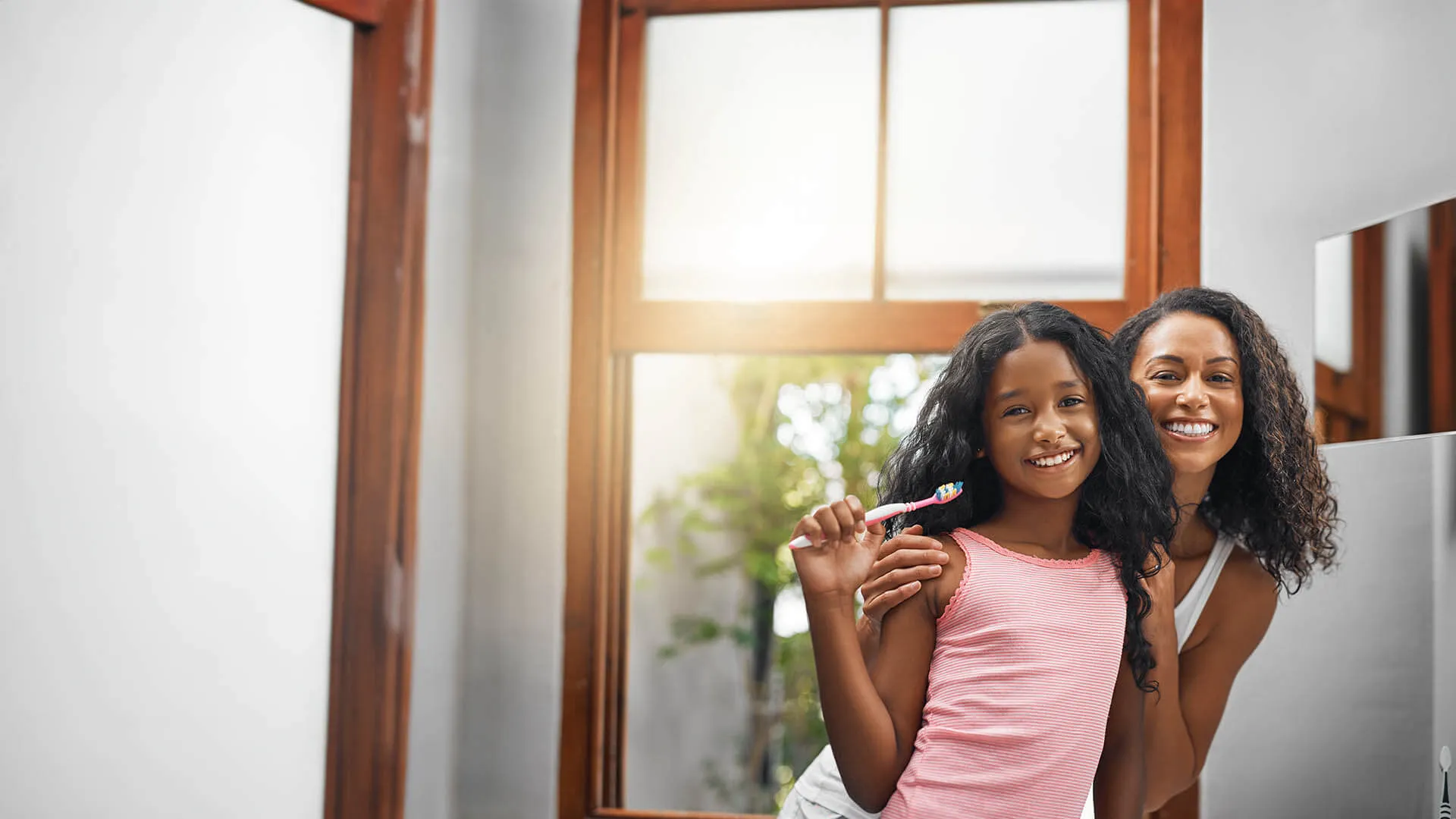 Smiling mother and daughter holding a toothbrush in a bright bathroom with wooden window frames.