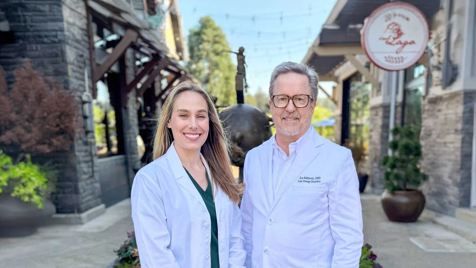 Two medical professionals in white coats standing outdoors in a sunny urban setting, smiling at the camera.