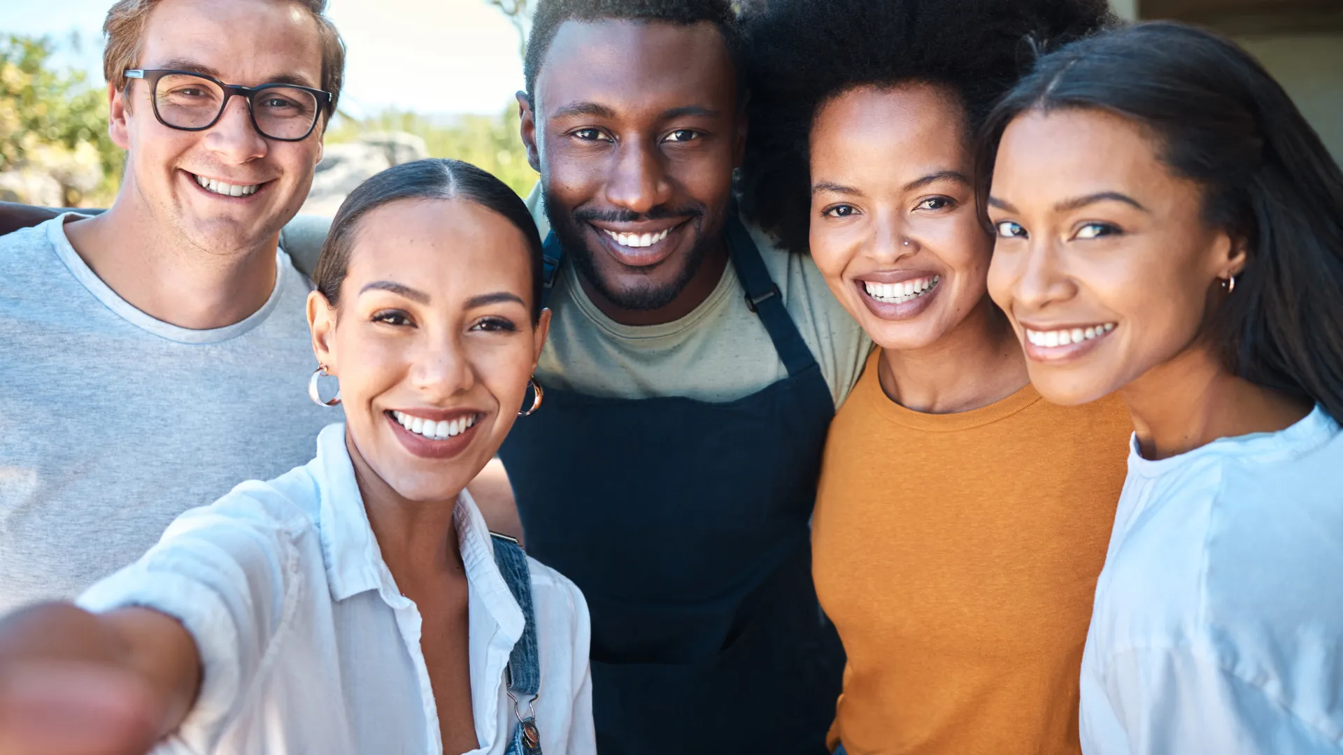 Group of five diverse friends smiling and taking a selfie outdoors on a sunny day