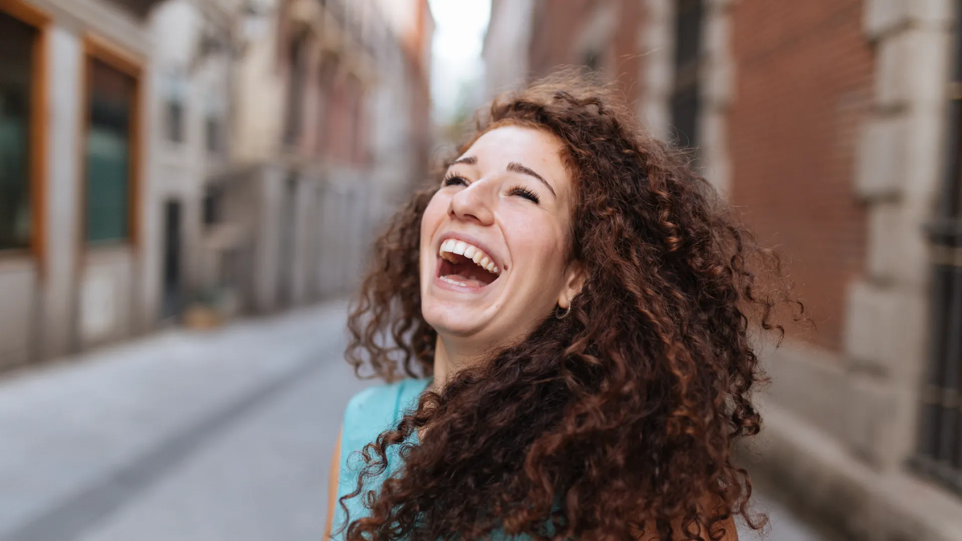 Joyful woman with curly hair laughing outdoors on a city street wearing a turquoise sleeveless top
