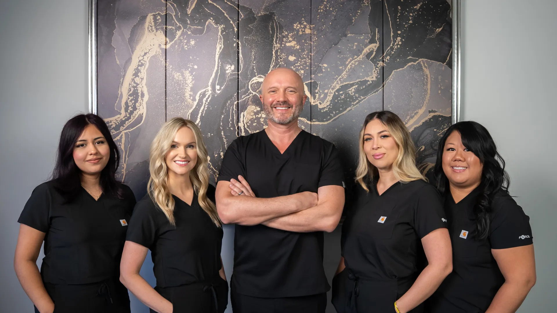 Smiling diverse medical team in black scrubs standing confidently against an artistic wall background.