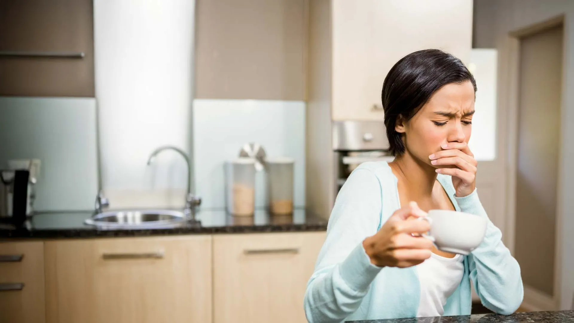 Woman in kitchen holding a cup and covering her mouth, appearing to cough or feel unwell.