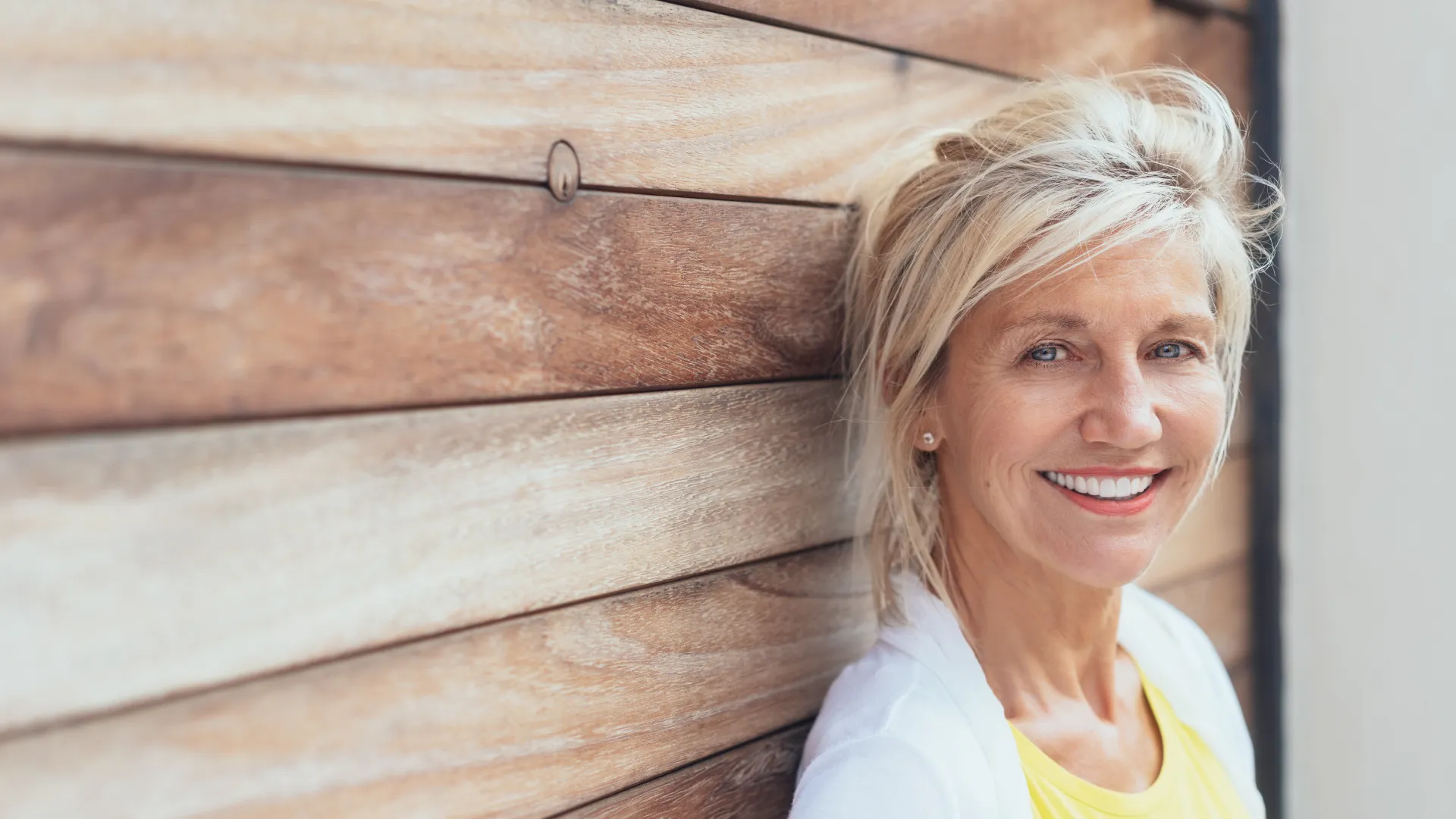 Smiling mature woman with blonde hair leans against a wooden wall wearing a yellow shirt and white cardigan.