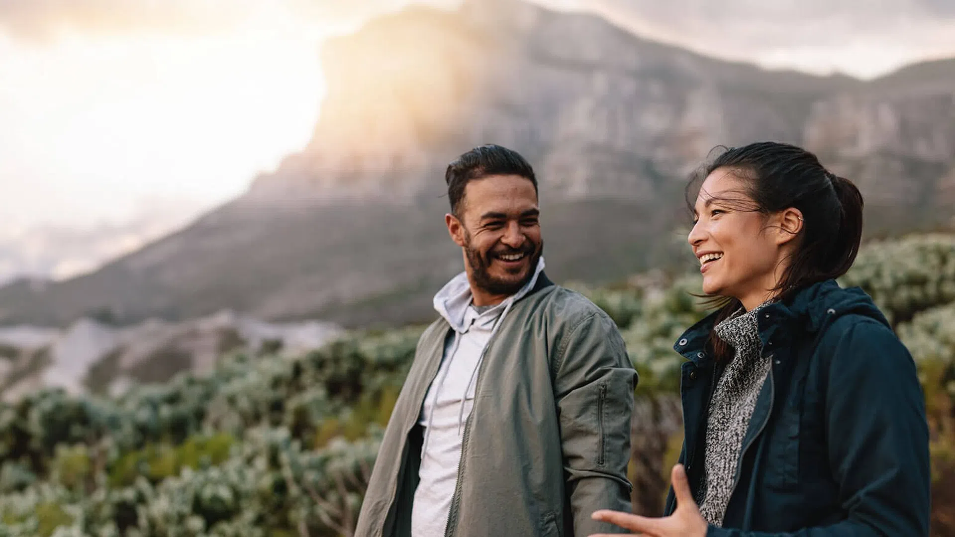 Smiling man and woman hiking outdoors in mountainous landscape during sunset with clouded sky.