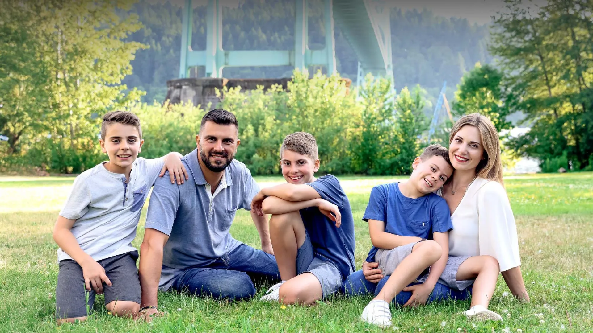 A happy family sitting on grass, smiling together, surrounded by greenery and under a bridge.