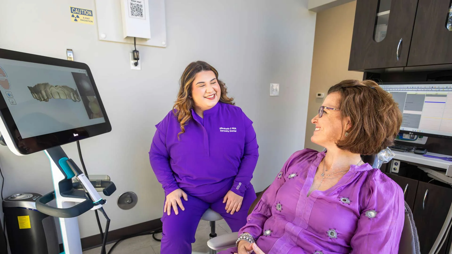 Dentist in purple scrubs consulting a smiling female patient in dental office with 3D dental imaging screen.