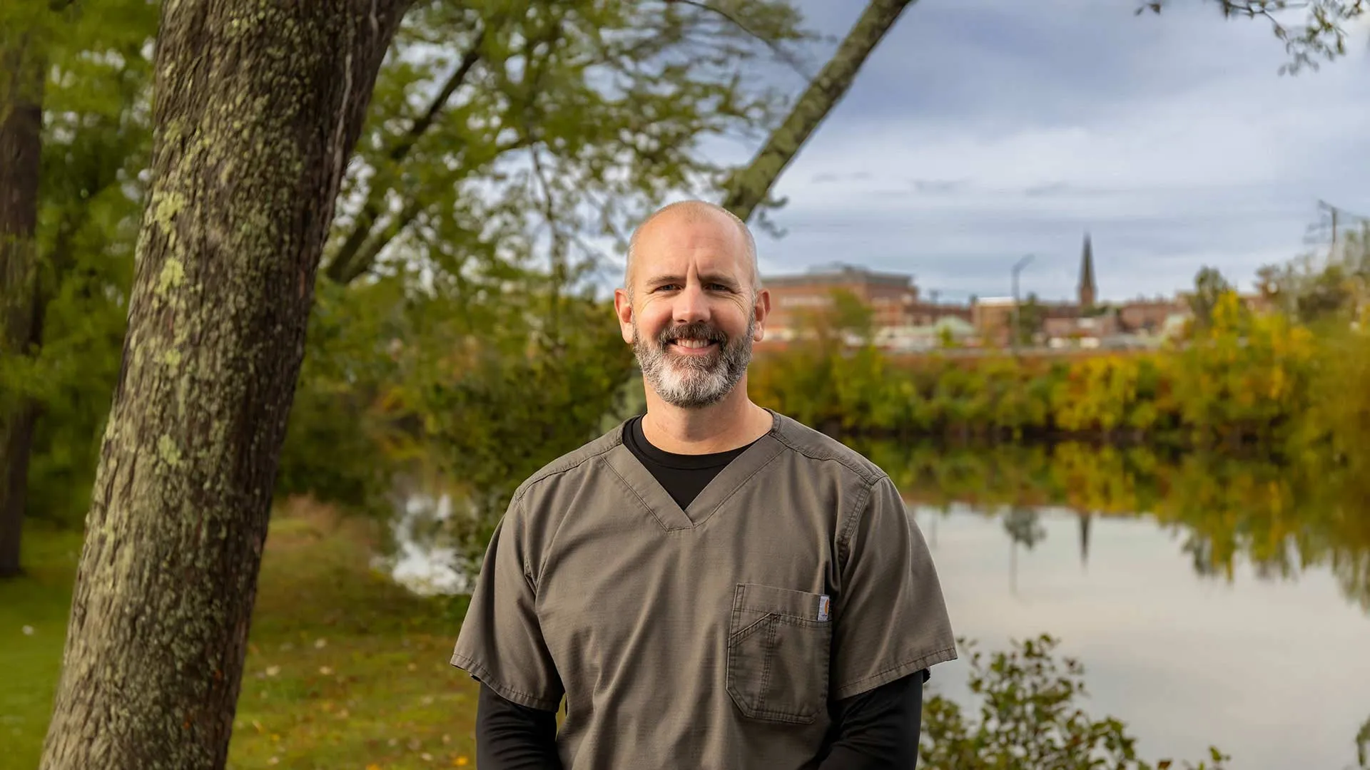 Smiling man in grey scrubs standing outdoors by a calm river with trees and a city skyline in the background