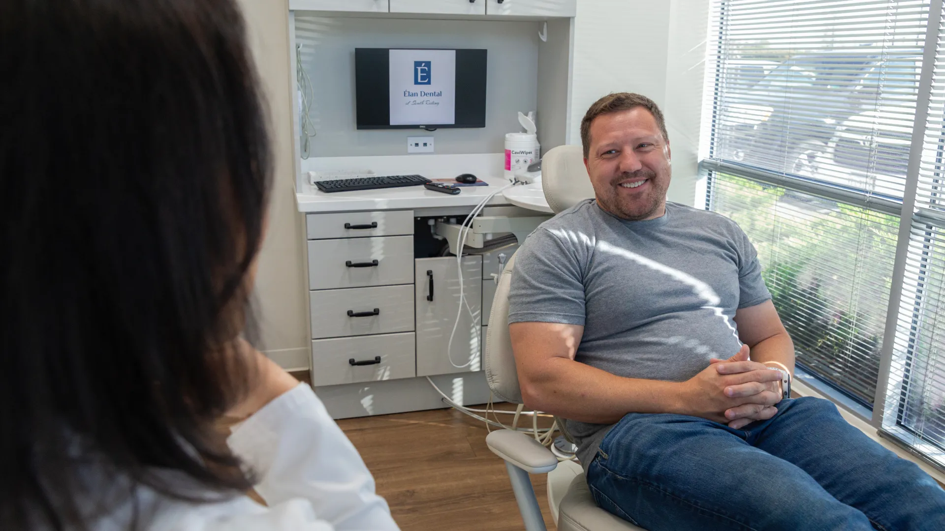 Man smiling while sitting in dental office chair talking to female dentist in a well-lit room.