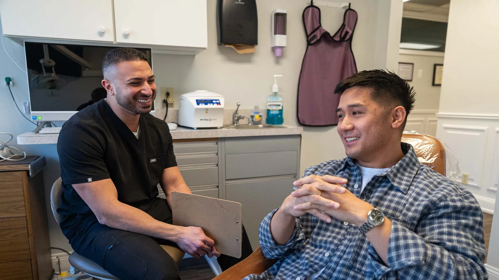 Dentist in black scrubs consulting with a smiling male patient sitting in a dental chair in a clinic.