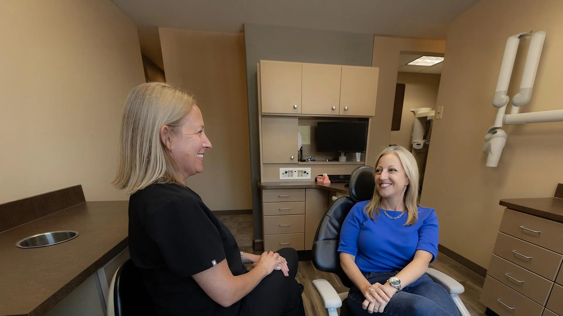 Dentist in black scrubs talking with patient seated in dental chair inside a modern clinic room.