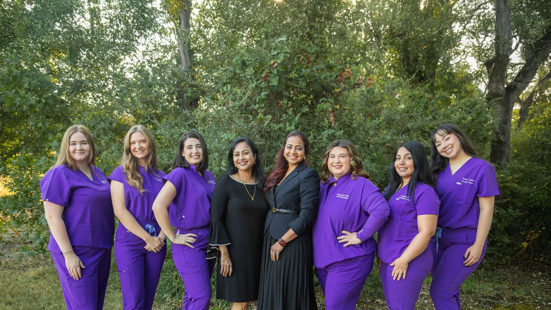 Group of eight women, five in purple scrubs and three in black dresses, standing outdoors in a green wooded area.