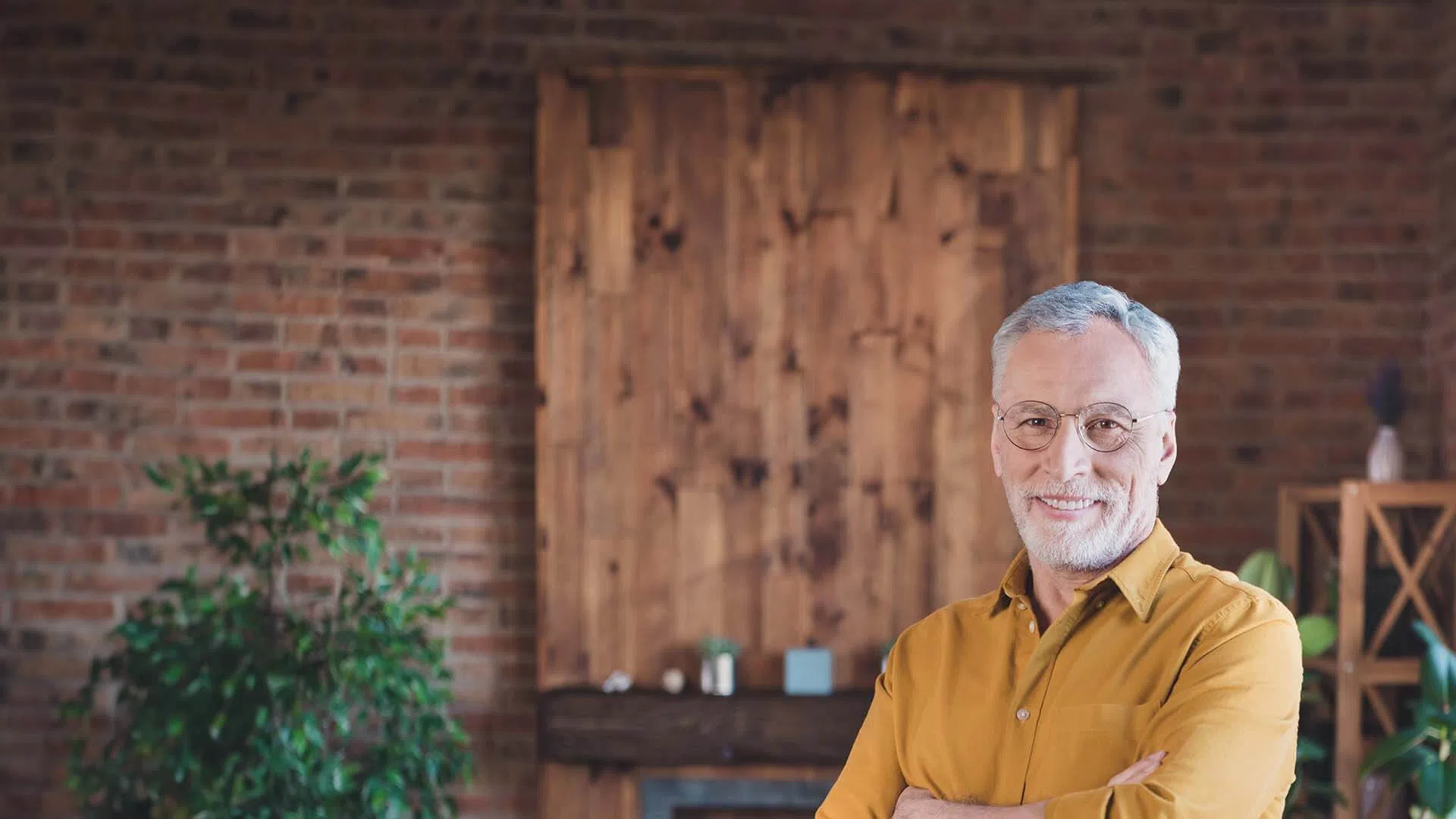 Smiling elderly man with gray hair and glasses wearing a mustard shirt standing in a cozy room with wooden wall and plants.
