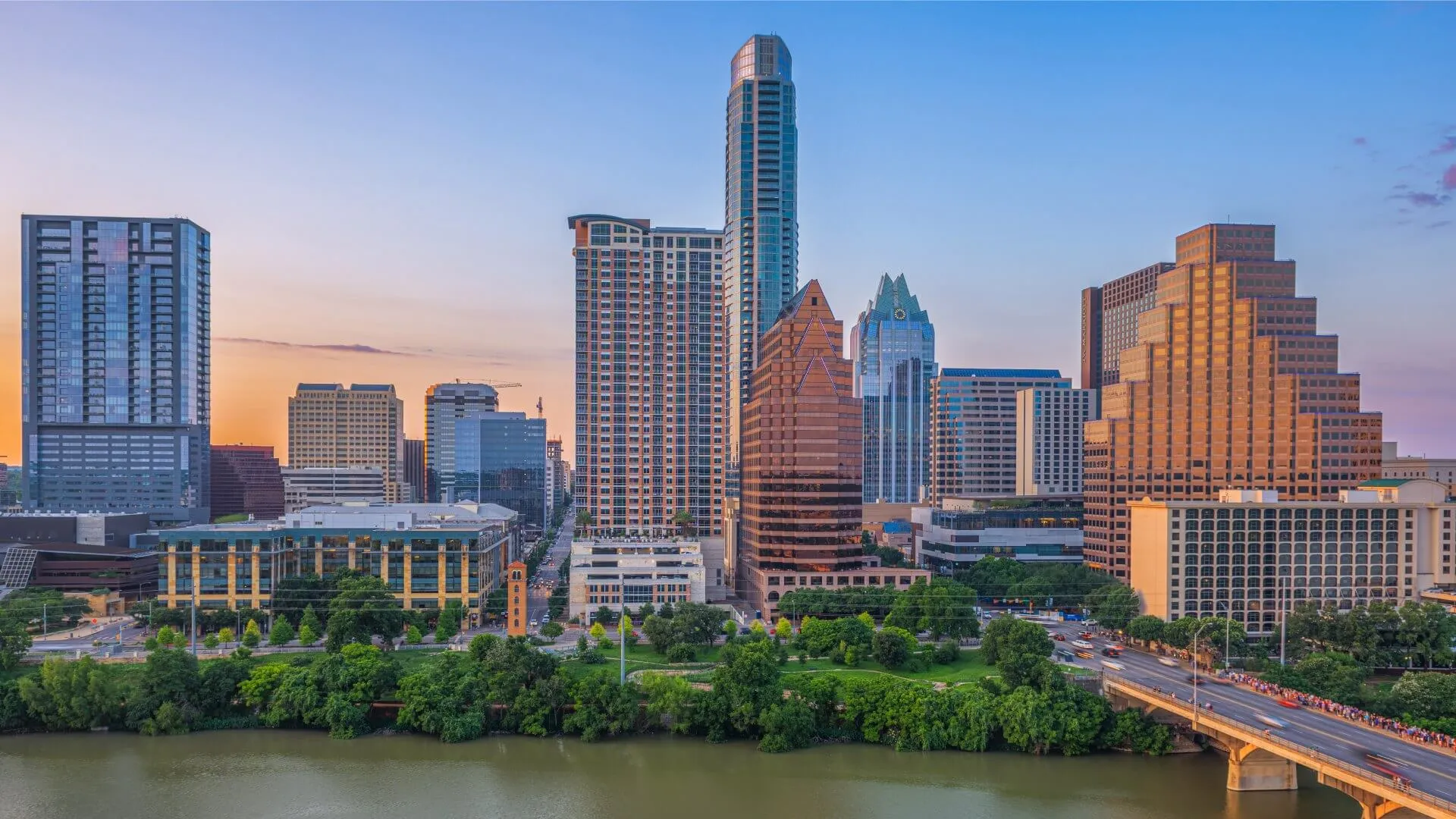 Austin city skyline at sunset featuring the Colorado River, green trees, and modern buildings with clear sky.