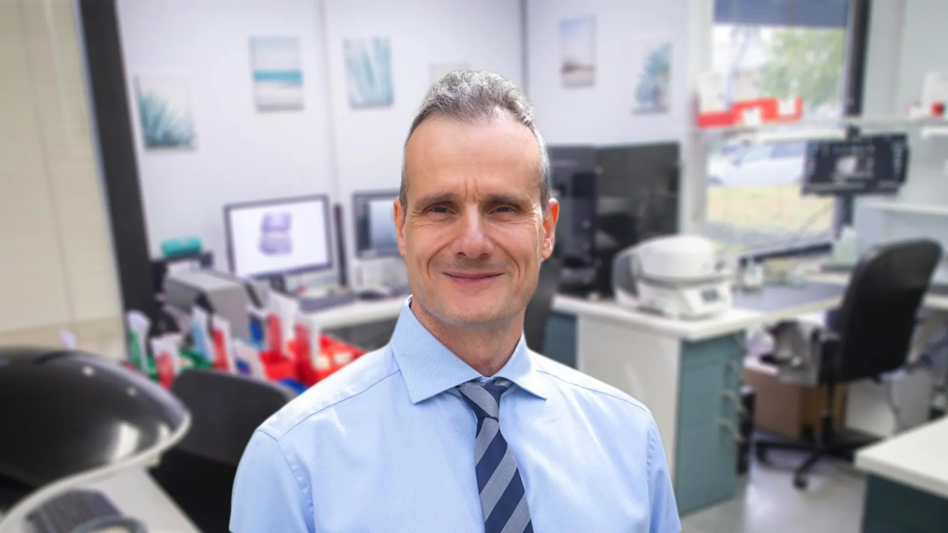 Professional man in a light blue shirt smiling in a modern office environment with computers and medical equipment.