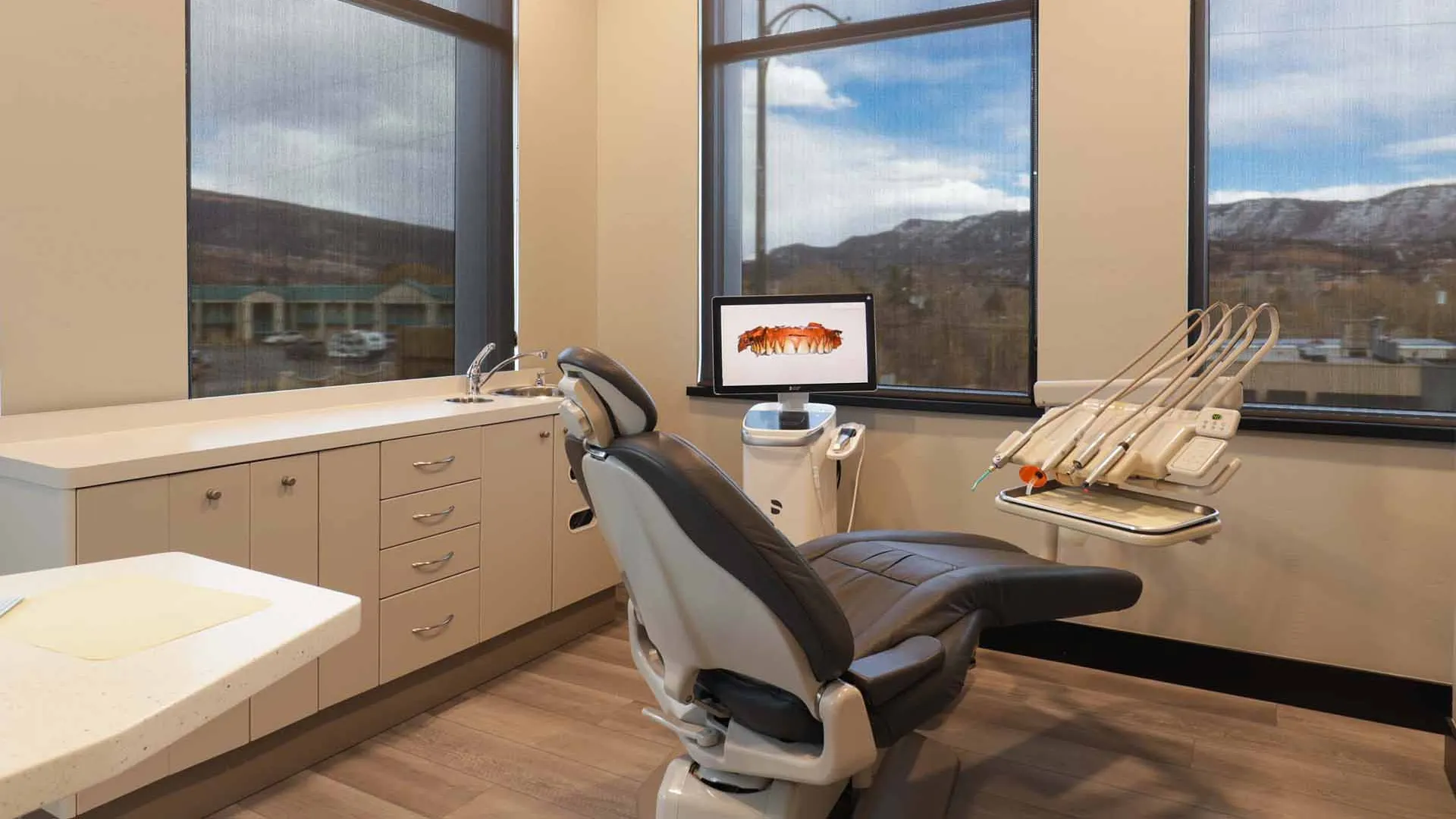 Modern dental exam room with reclining chair, dental tools, computer screen, and large windows showing mountainous view.