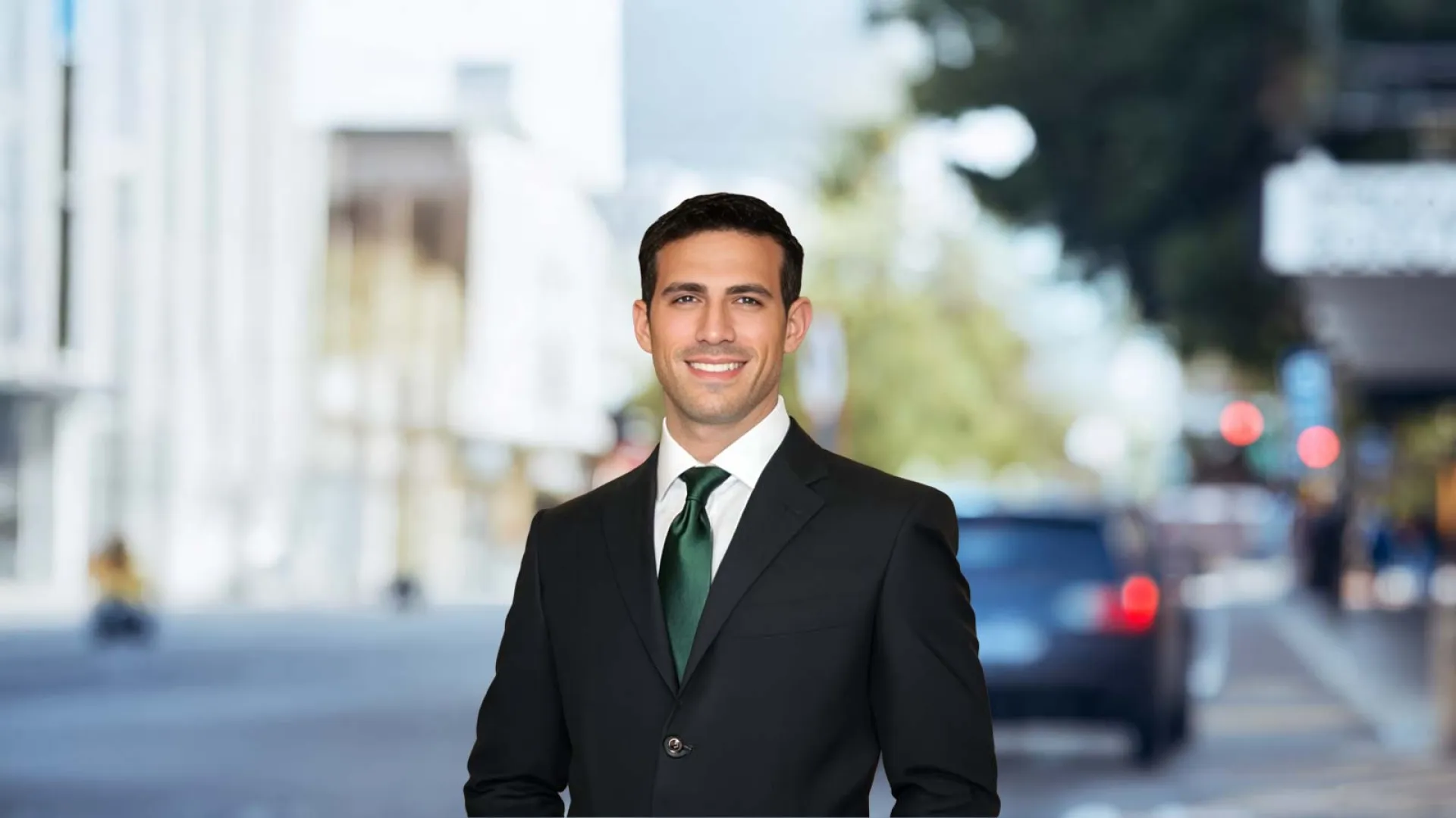 Confident young businessman in black suit and green tie smiling on busy city street in daylight.