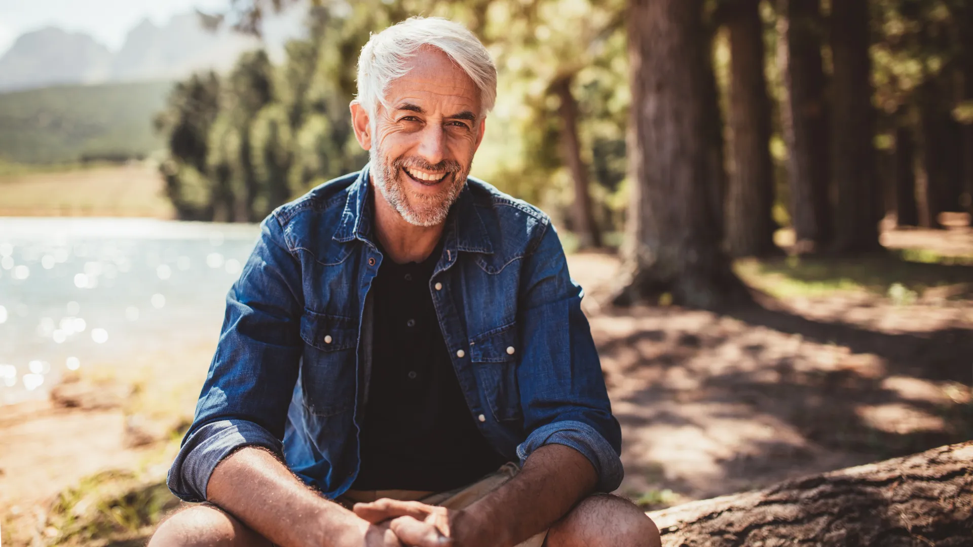 Smiling senior man sitting outdoors by a lake wearing a denim jacket on a sunny day