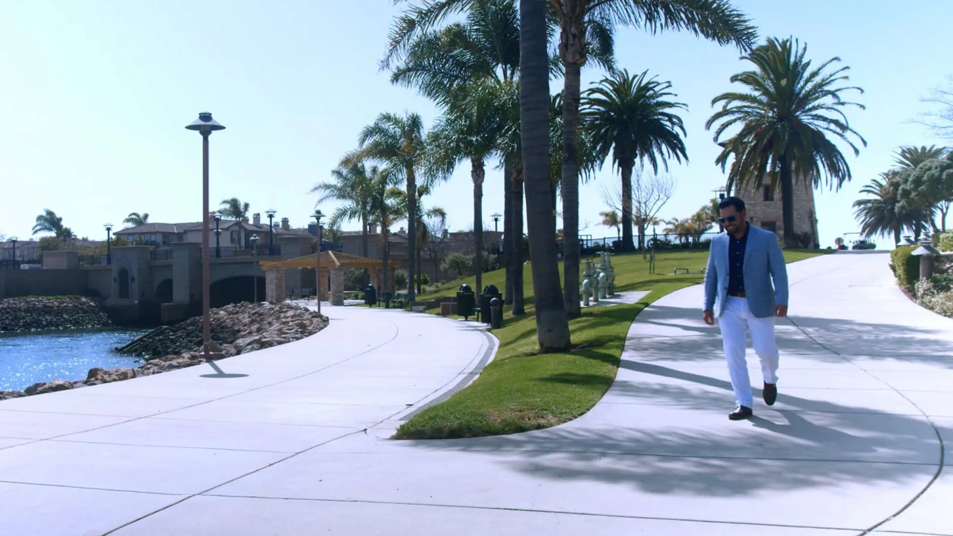 A man walks along a palm-lined pathway by a serene waterfront with lush greenery.