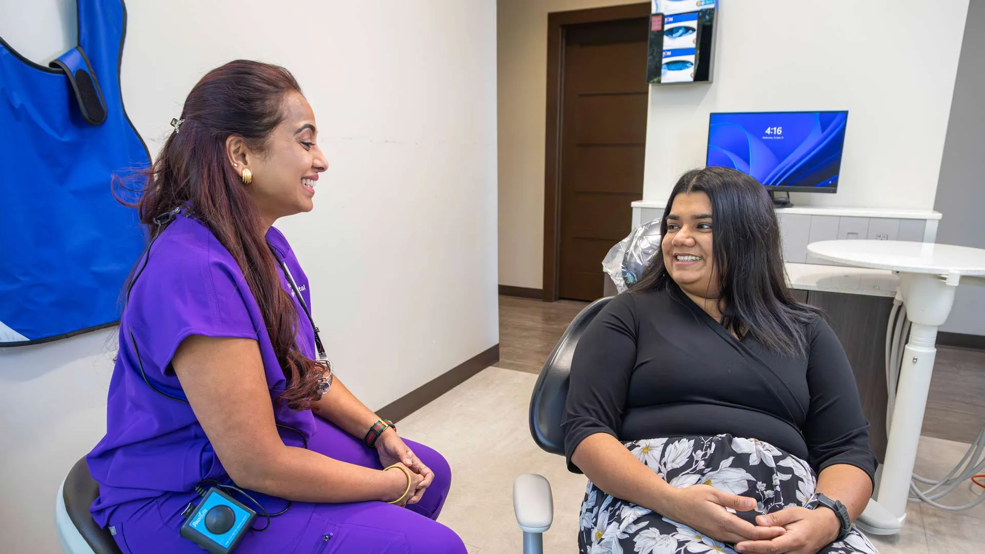 Dentist in purple scrubs talking and smiling with female patient in dental office chair