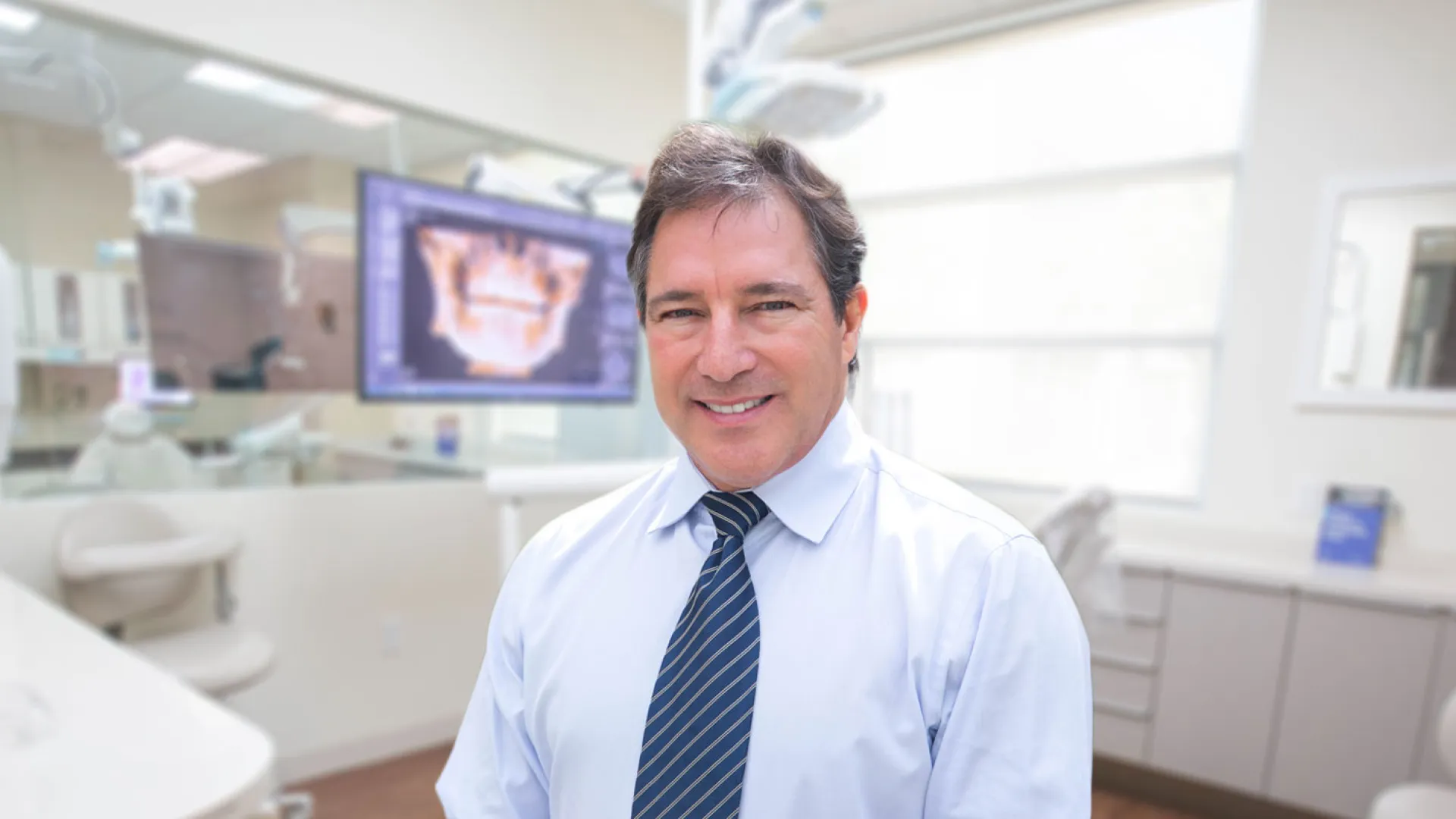 Smiling dentist in a modern dental office with equipment and a patient monitor in background.