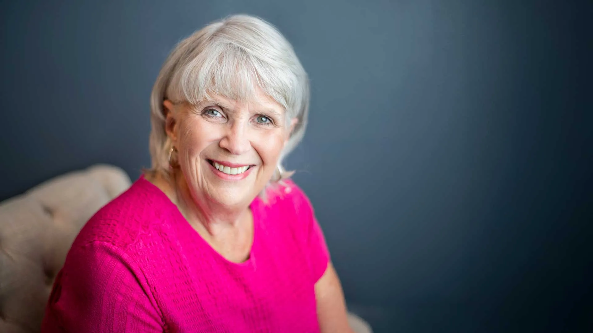 Smiling elderly woman with short white hair wearing a bright pink top against a blue background