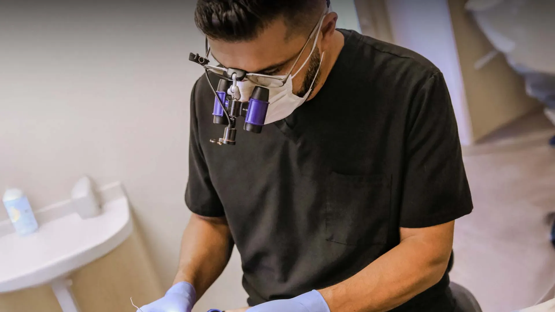 A dentist in scrubs performing an oral procedure with magnifying glasses.