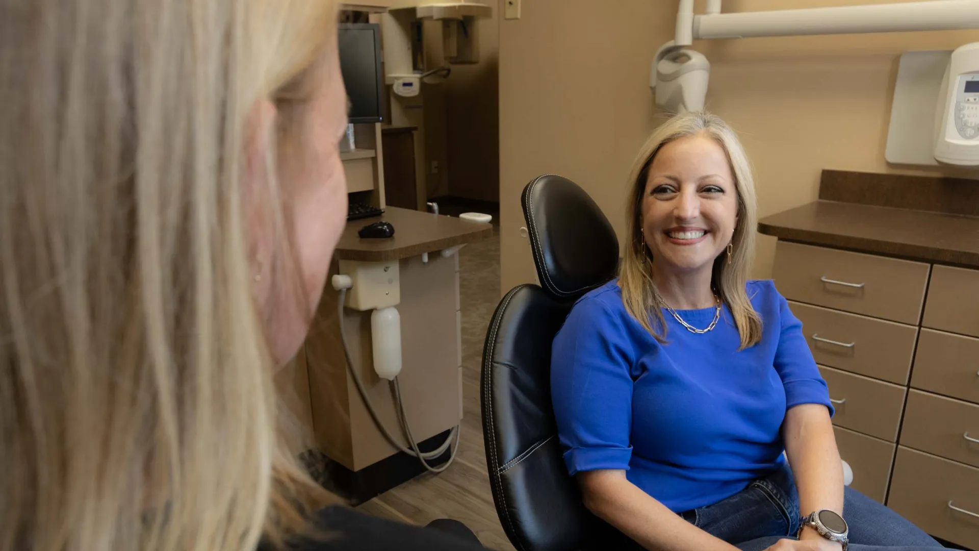 Smiling woman in blue shirt sitting in dental chair talking to another person in dental office.