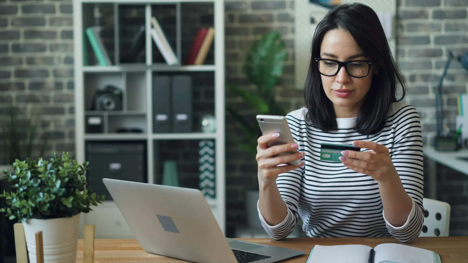 Woman wearing glasses using smartphone and credit card for online shopping at a desk with laptop and notebook.