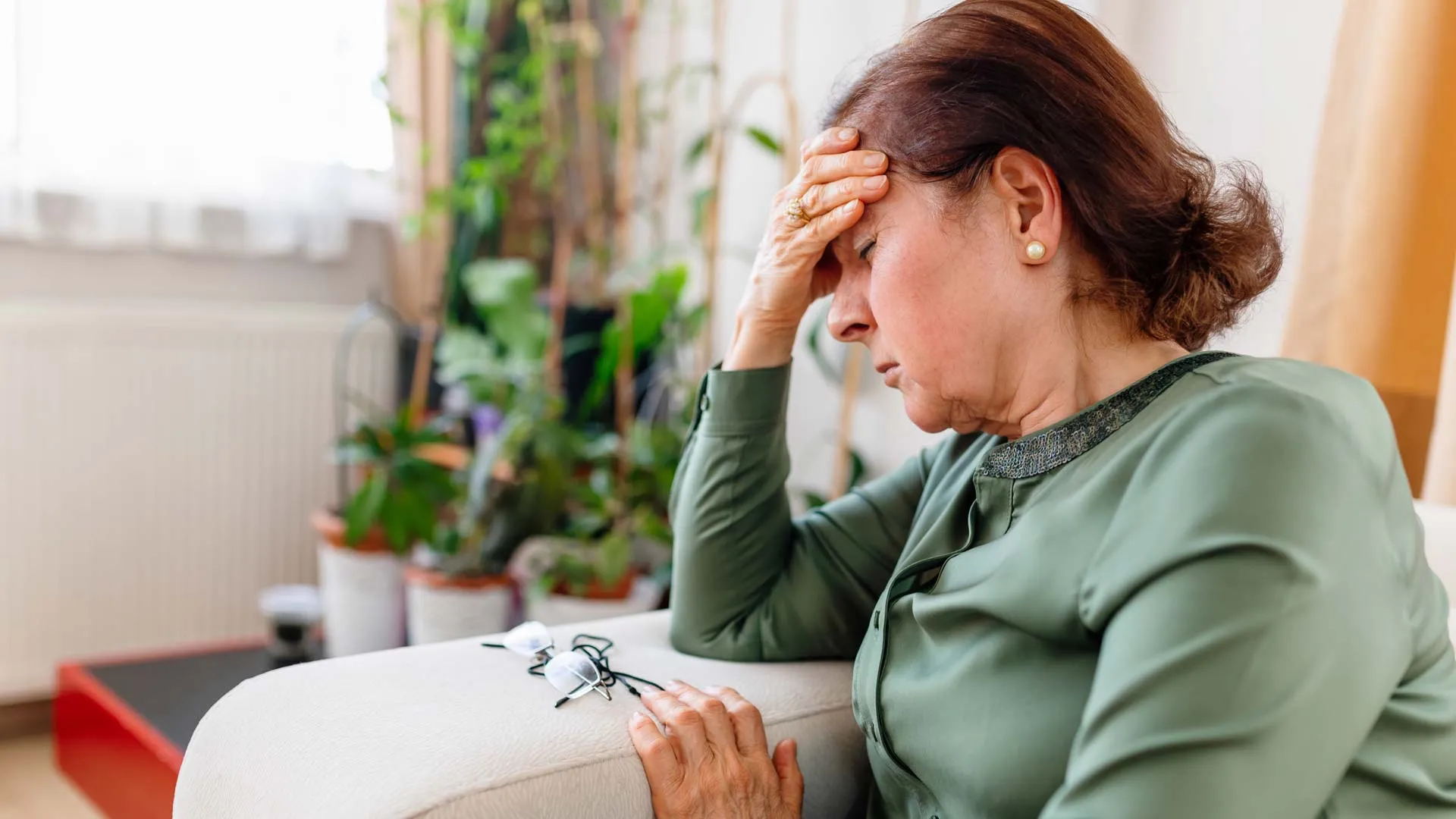 Middle-aged woman in green blouse sitting on couch, holding forehead, headache