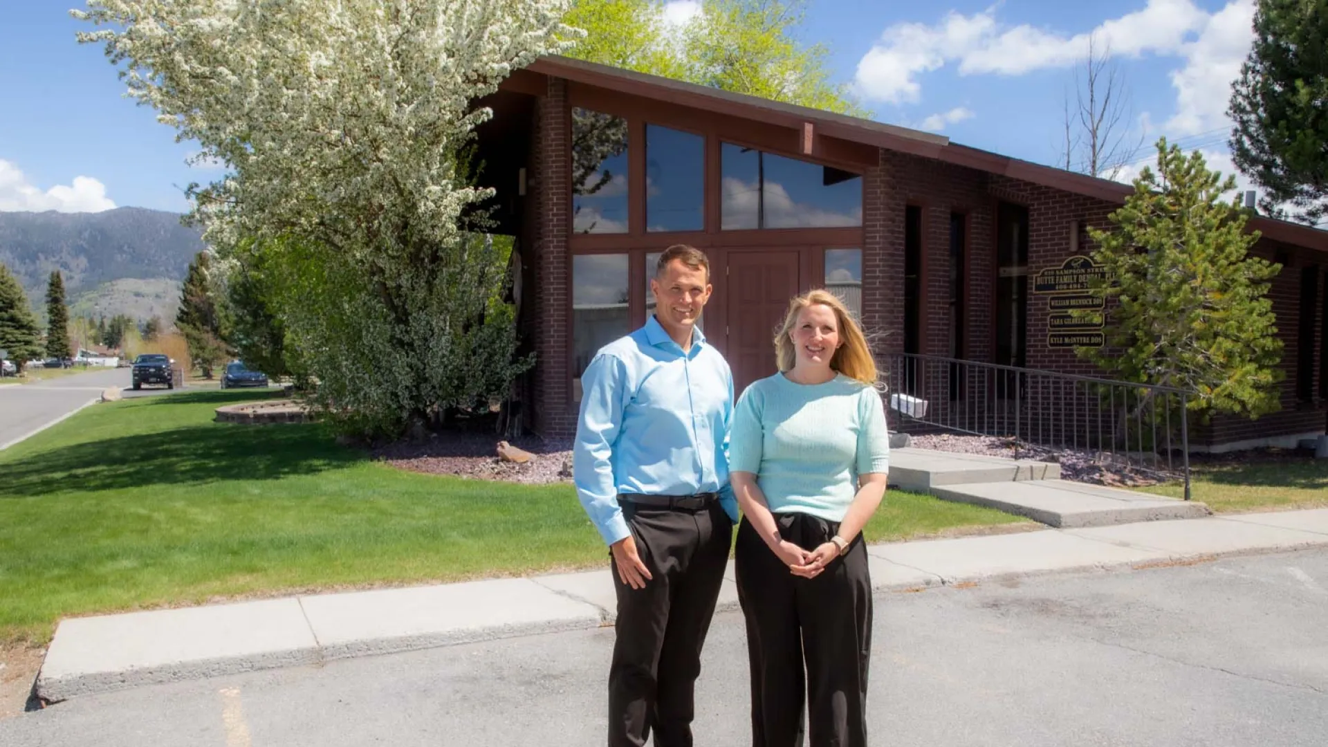 Man and woman standing outside a brick office building with trees and mountains in the background on a sunny day
