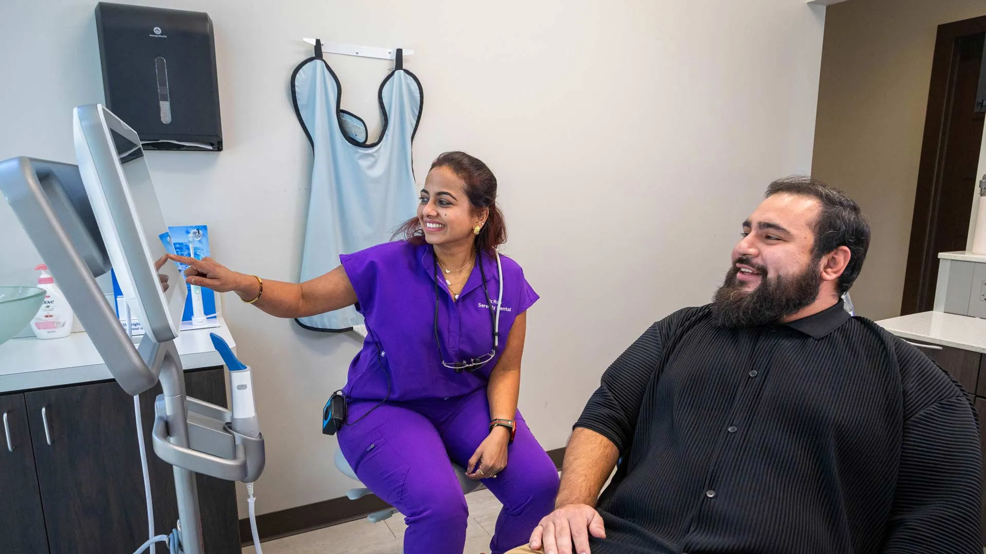 Female medical professional in purple scrubs showing a patient information on a monitor in a clinic room.