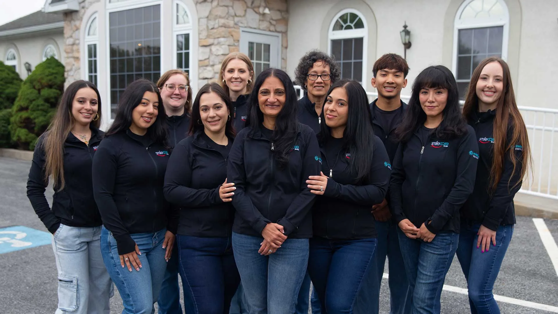 Diverse group of smiling coworkers wearing black jackets standing outside a building in a parking lot.