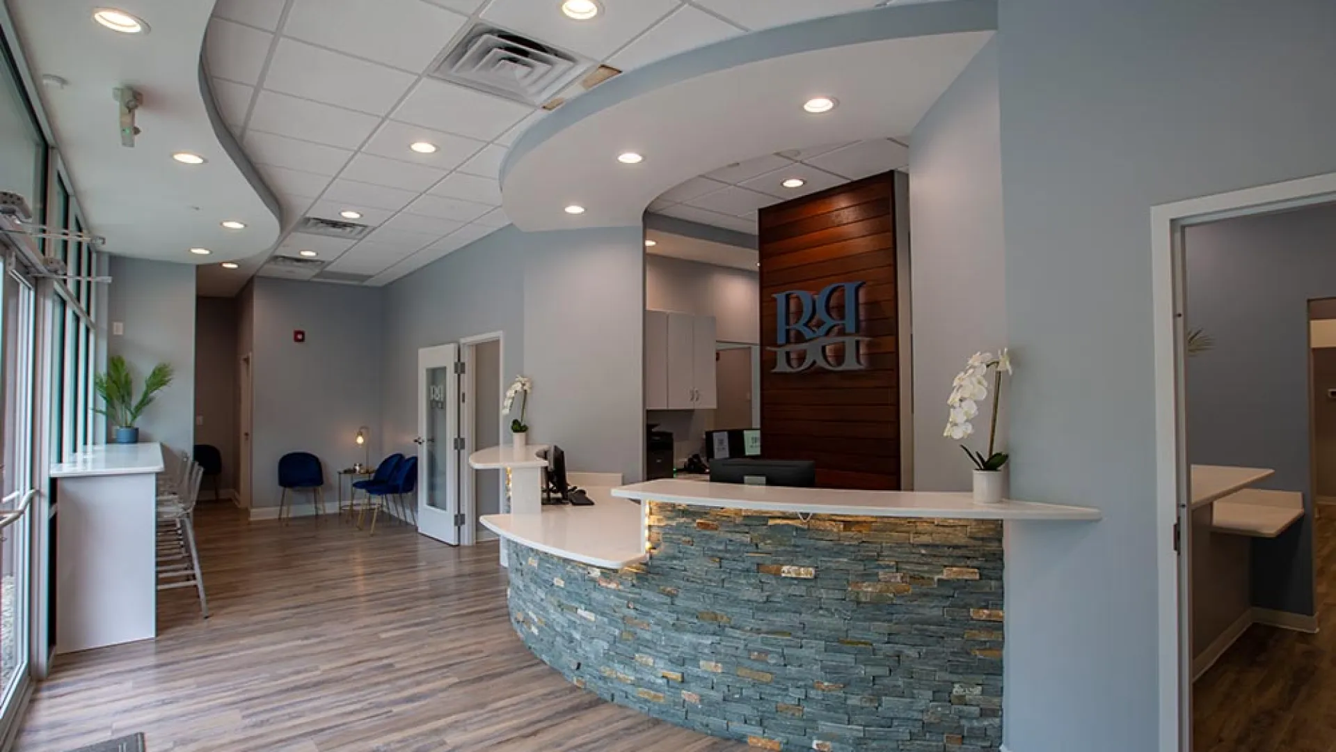 Modern dental office reception area with curved stone desk, wood flooring, blue chairs, and soft lighting.
