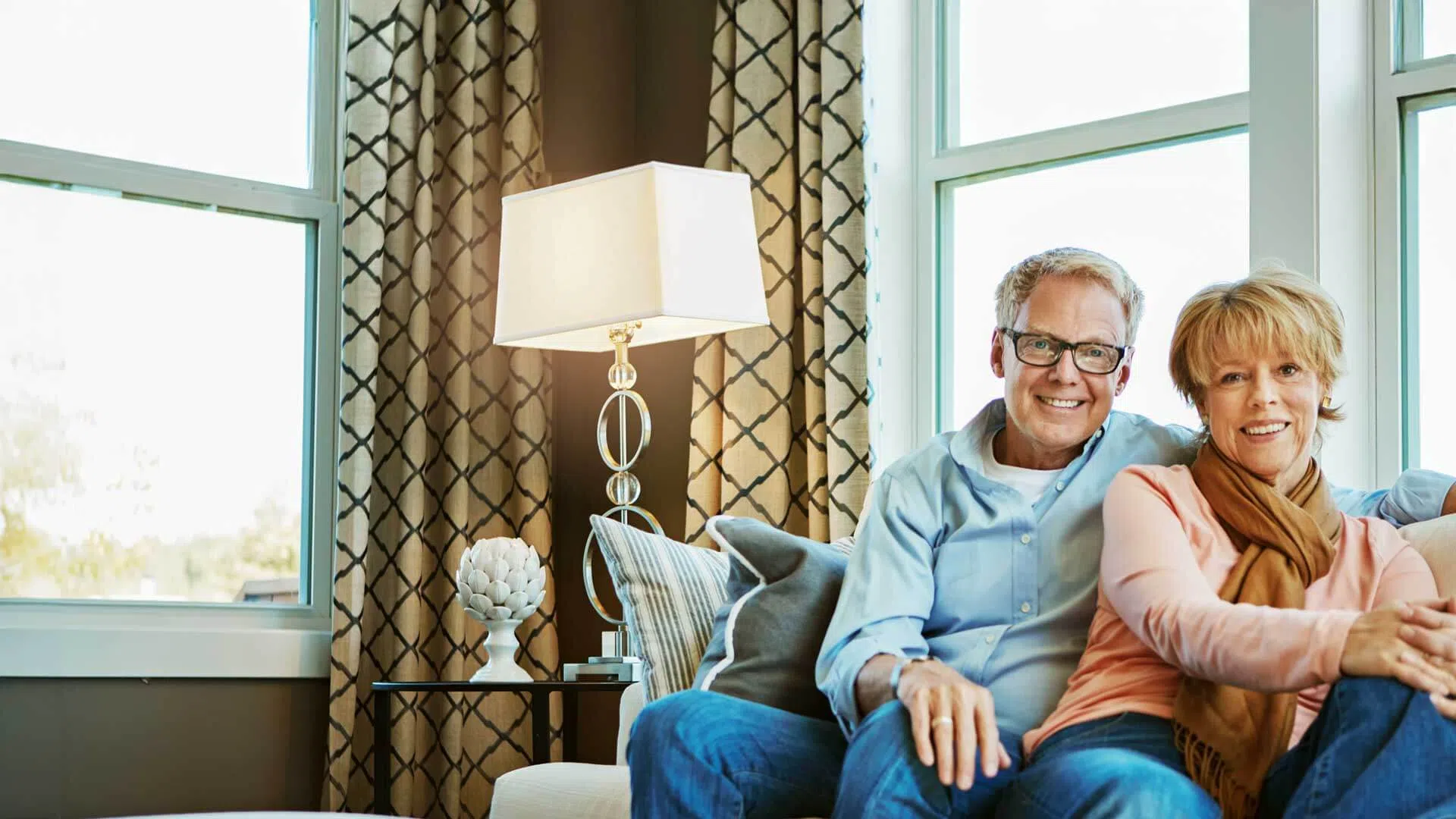Happy older couple sitting together on a cozy couch in a bright living room with a lamp and patterned curtains