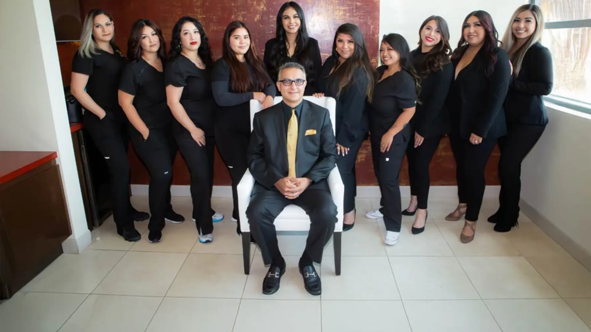 A professional team of women in black outfits posing with a man in a suit in a modern office setting.