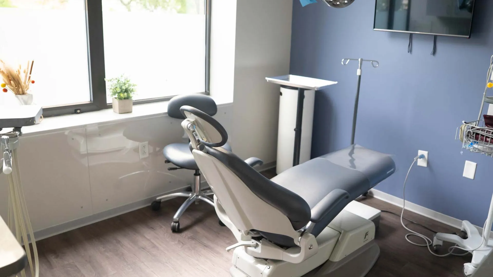 Modern dental office with an adjustable chair, stool, medical equipment, and natural light from a large window.