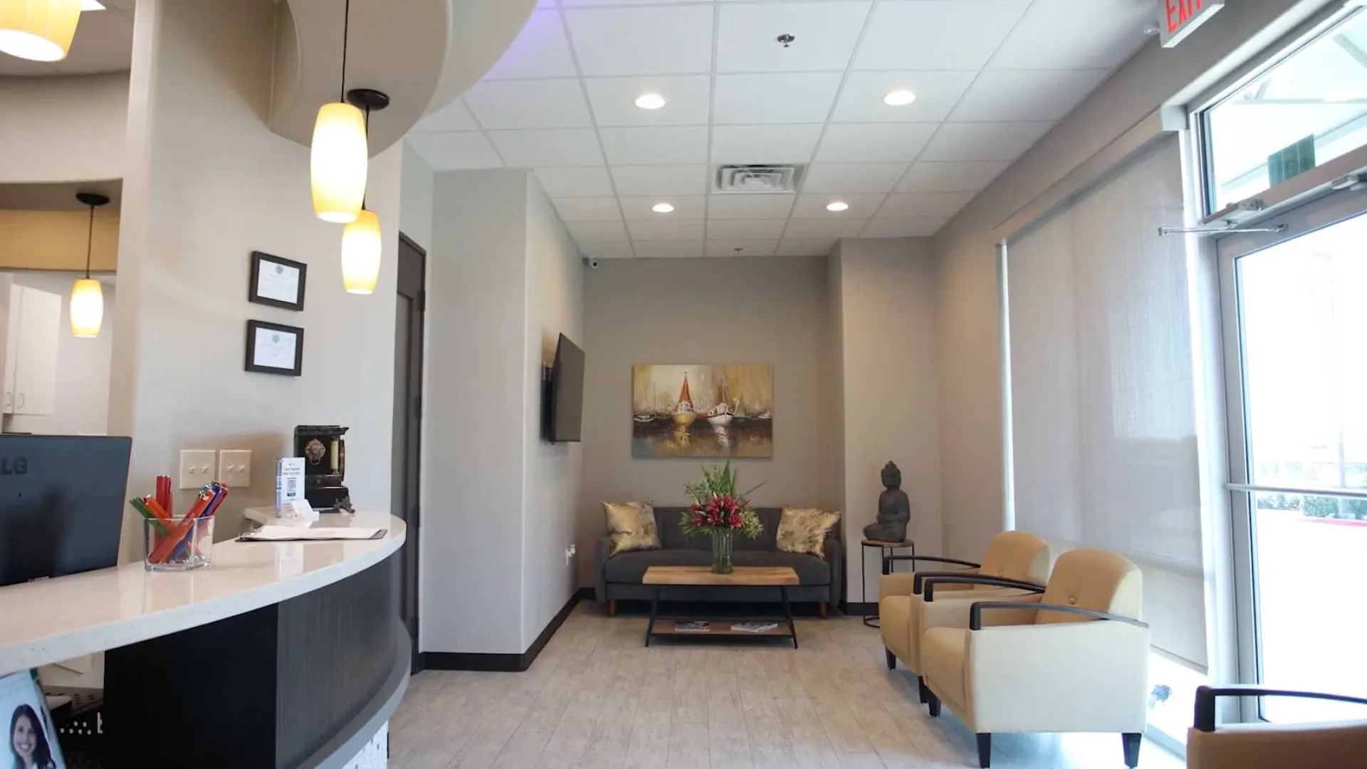 Modern waiting room with beige chairs, floral centerpiece, artwork, and curved reception desk with hanging lights.