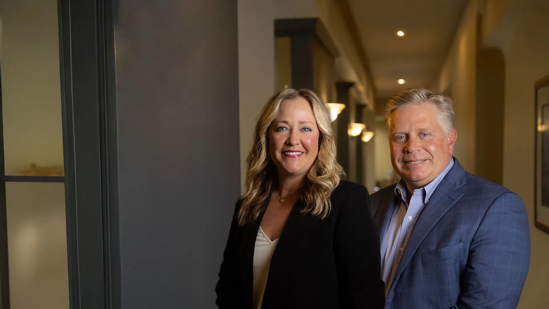 Professional middle-aged man and woman smiling in an office hallway with warm lighting.
