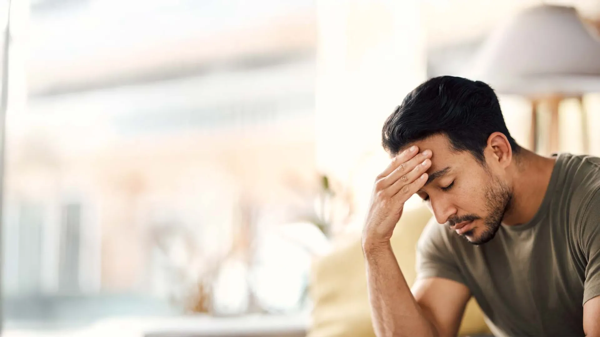 Man sitting with hand on forehead, appearing stressed or worried indoors with blurred background.