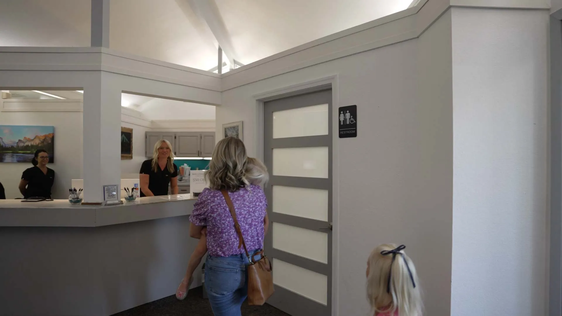 Mother carrying child walking toward reception desk with two women behind counter in bright office interior