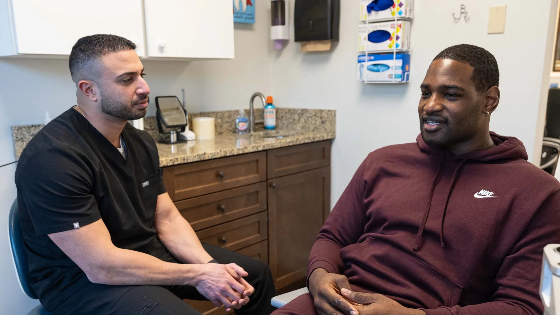 Dentist in black scrubs consulting a male patient in a burgundy Nike hoodie in a modern dental office.