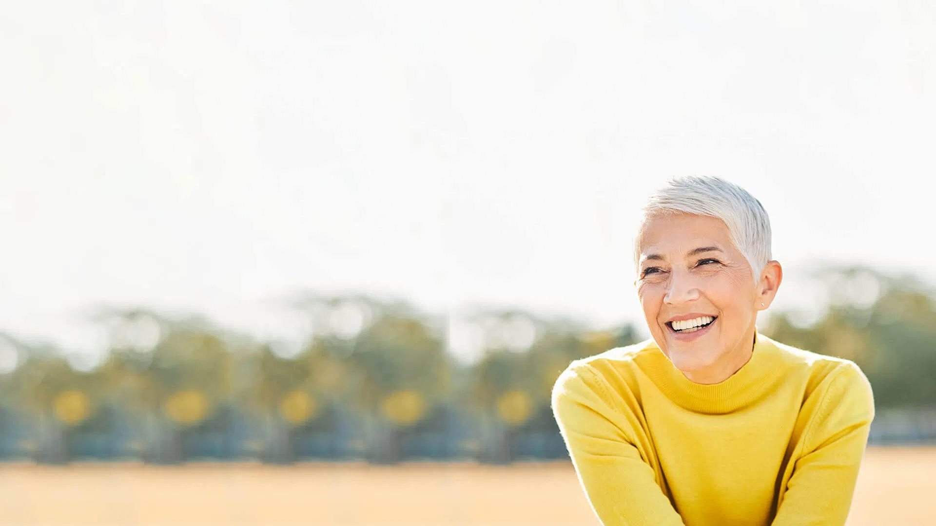 Smiling mature woman with short white hair wearing a bright yellow sweater outdoors with blurred trees background