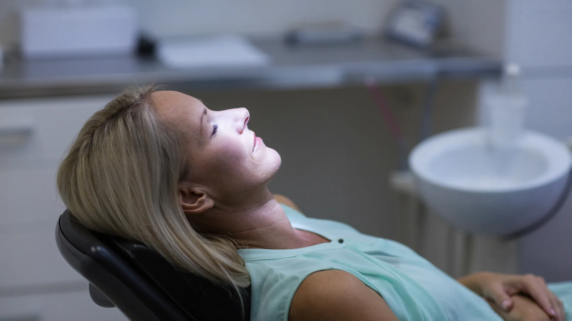 Woman reclining in dental chair with eyes closed, illuminated by bright dental light in clinic