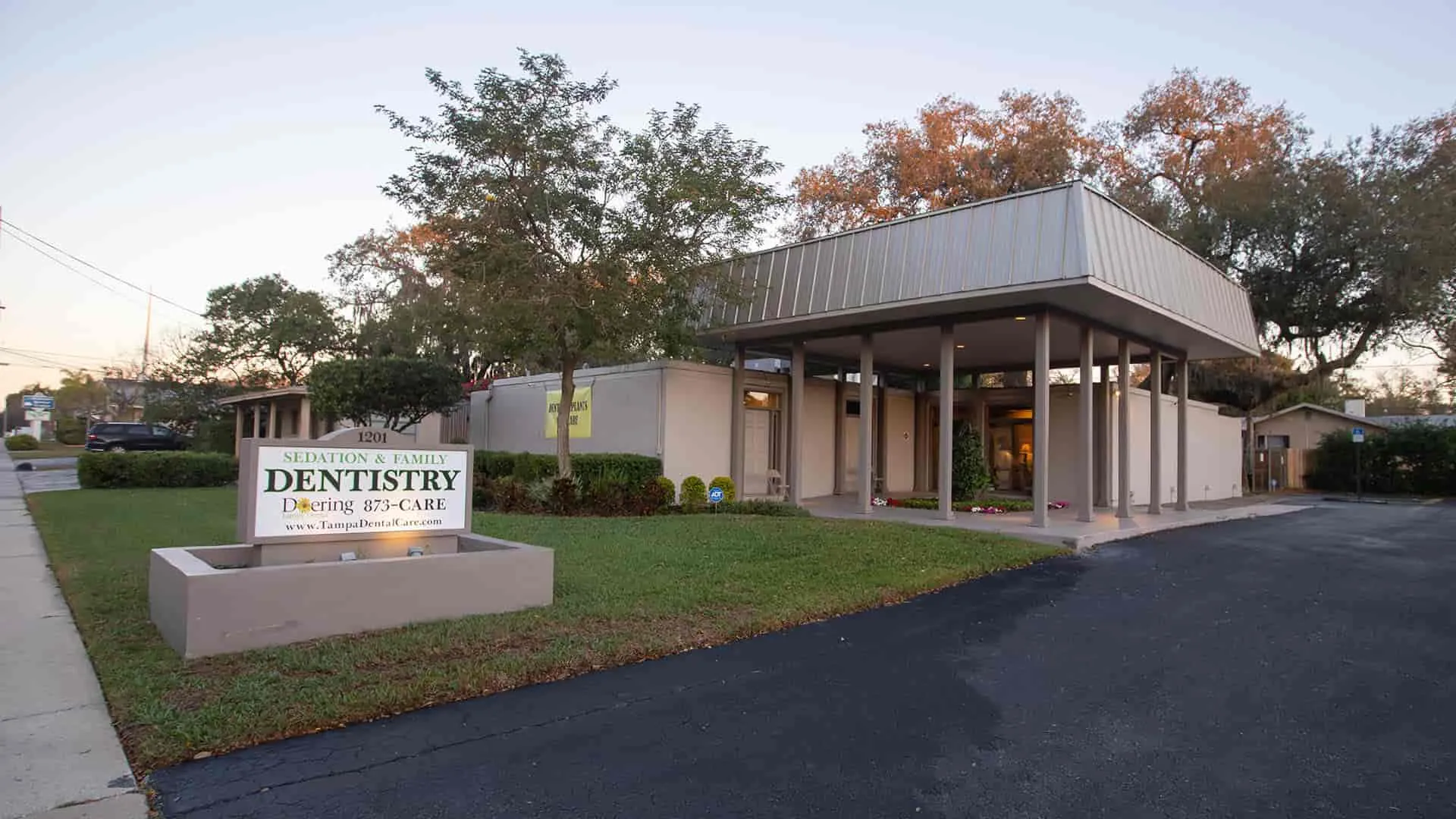Sedation and family dentistry clinic building with signage on a grassy lawn and surrounding trees.