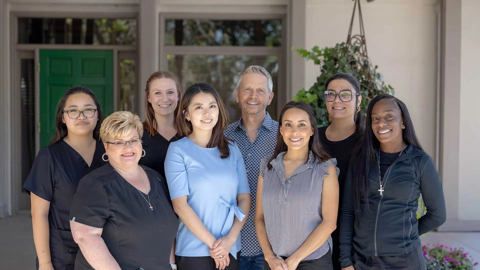 Diverse group of eight smiling adults standing outdoors in front of a building with a green door and potted plants.