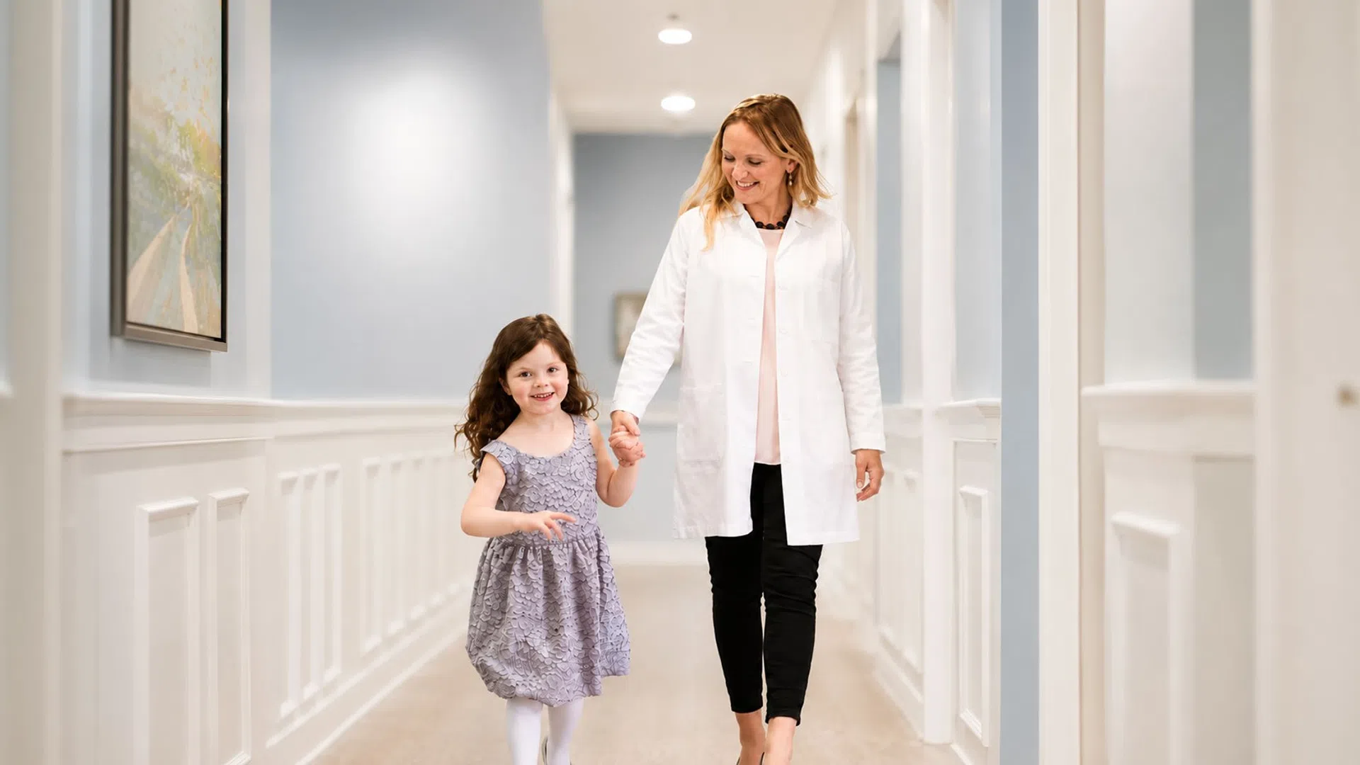 Female doctor holding hands with smiling young girl walking down a bright hallway in a medical clinic.