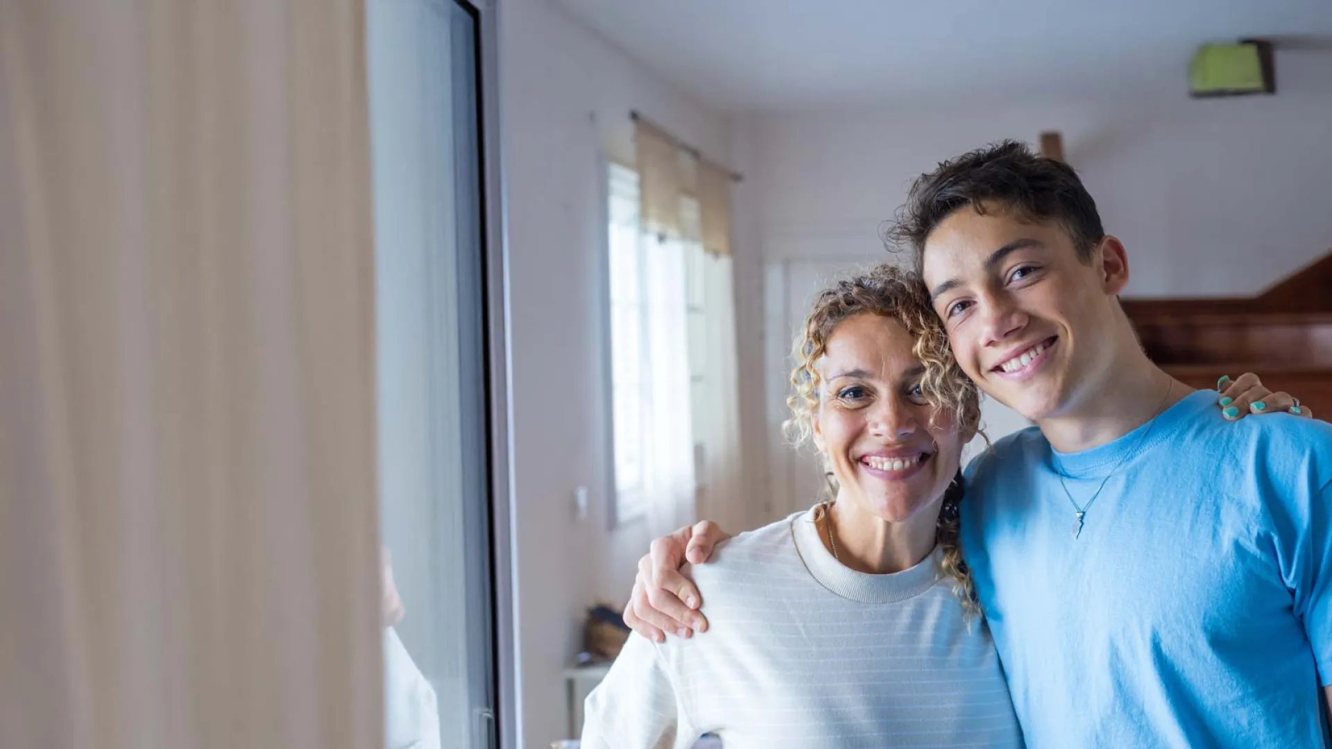 Smiling mother and teenage son embracing inside a bright home by a window with natural light.